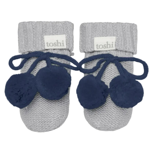 Pair of baby booties with navy pom-poms and gray cuffs on a white background, featuring the brand 'toshi'.